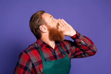 Photo of excited man gardener wear red plaid shirt green apron look up empty space tell say speak isolated on purple color backgroundの写真素材