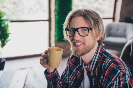 Portrait photo of young successful data science specialist programmer man in glasses holding cup hot coffee enjoying morning routine at officeの写真素材
