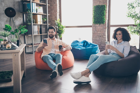 Full body portrait of two young corporate workers sit beanbag use smart phone modern loft interior office indoorsの写真素材