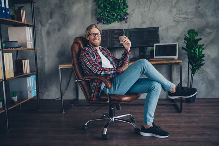 Photo of blond hair young businessman own startup data science sitting armchair near workstation with computer drinking takeaway coffee at officeの写真素材