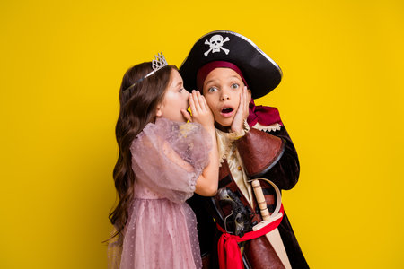Portrait of two small kids halloween pirate princess costume whisper ear isolated on yellow color backgroundの写真素材