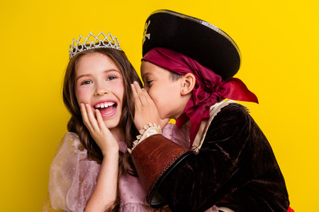 Photo of two kids halloween princess pirate costume whisper tell ear isolated on yellow color backgroundの写真素材
