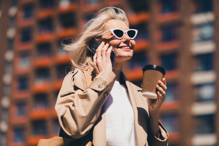 Portrait of happy retired woman smile spring good mood sunglass talk phone coffee weekend wear beige coat outdoors outside park streetの写真素材