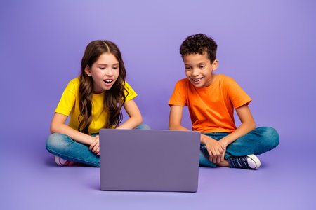 Full length photo of lovely two little kids netbook eshopping dressed stylish orange yellow garment isolated on purple color backgroundの写真素材