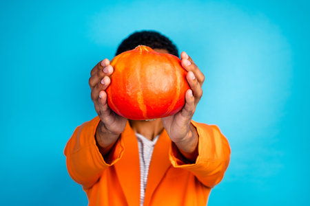 Photo of attractive guy showing in camera pumpkin vegetable for thanksgiving day hide face isolated blue color backgroundの写真素材