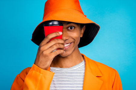 Portrait of happy guy smiling hide face with to go coffee mug wear stylish hat isolated blue color backgroundの写真素材