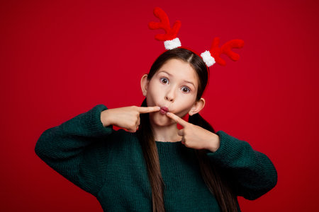 Photo of cute little girl fingers press cheeks fish lips wear pullover christmas festive time isolated on bright red color backgroundの写真素材