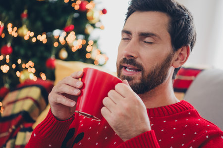 Photo of nice young man closed eyes drink coffee ugly sweater christmas time apartment indoorsの写真素材
