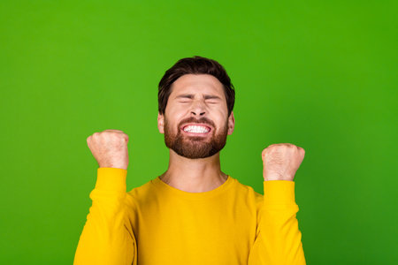 Photo of overjoyed man with stylish beard dressed yellow shirt clenching fists win gambling shout isolated on green color backgroundの写真素材