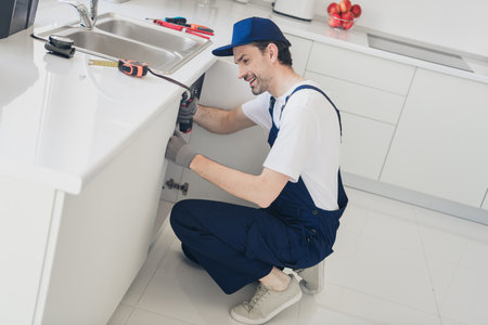 Skilled handyman in blue uniform repairing sink cabinet in modern kitchen with professional toolsの写真素材