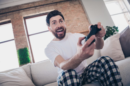 Photo of cheerful funny positive man wear pajamas sit on sofa play video game nice weekend indoors apartment flatの写真素材