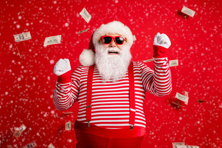 Photo of retired grandfather grey beard raise fists huge load money wear santa x-mas costume suspenders sunglass gloves striped shirt cap isolated red color backgroundの写真素材