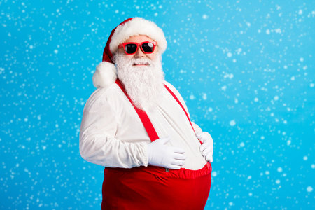 Photo of crazy overweight santa claus touch his big belly enjoy x-mas christmas newyear tradition feast wear cap suspenders overalls isolated over blue color backgroundの写真素材