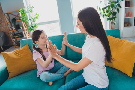 Cheerful positive charming mum and her lovely nice small daughter sitting on sofa clapping palms playing indoorsの写真素材