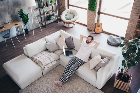 Photo of relaxed attractive man wearing nightwear hold arms behind head sit on divan weekend vacation loft interior indoorsの写真素材