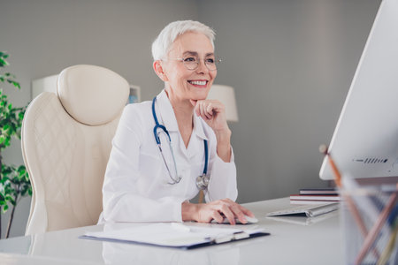 Positive lovely mature lady doctor working computer online consultation recipe wear white lab coat light hospital officeの写真素材