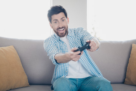 Photo of handsome bearded alone mature age adult man staying home to play favorite console holding joystick in living room with huge couchの写真素材
