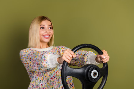 Excited young woman holding steering wheel, ready for adventure, wearing colorful knit sweater against khaki backgroundの写真素材