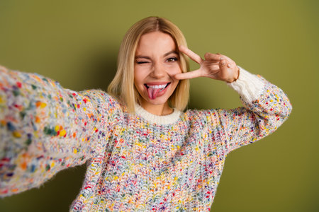 Cheerful young woman in colorful knitwear making peace sign and puckering lips against khaki backgroundの写真素材