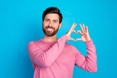 Smiling man in a pink sweater making a heart shape with his hands on a blue background, expressing...
