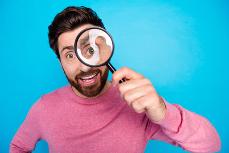 Joyful man with beard and pink sweater playfully looking through magnifying glass on bright blue...