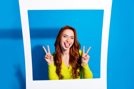 Photo of lovely young lady instant photo frame stick tongue out v-sign dressed stylish yellow garment isolated on blue color backgroundの写真素材