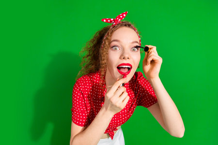 Photo portrait of pretty young girl apply pomade mascara wear trendy red retro outfit isolated on green color backgroundの写真素材