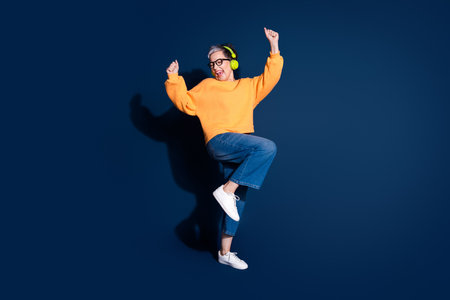 Full body photo of attractive mature woman dance have fun headphones dressed stylish yellow clothes isolated on dark blue color backgroundの写真素材