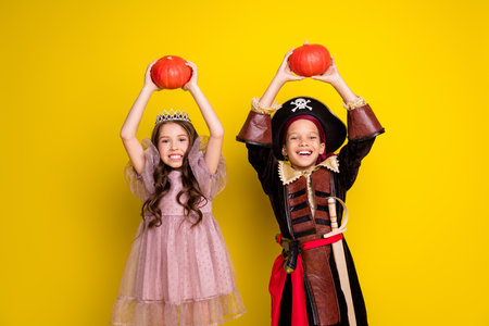 Portrait of two kids halloween princess pirate costume hold pumpkin above head isolated on yellow color backgroundの写真素材