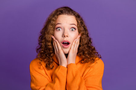 Portrait of wavy red hair young surprised woman in orange stylish turtleneck touch cheeks shocked isolated on violet color backgroundの写真素材
