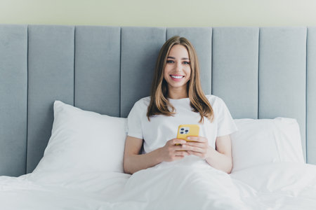 Photo of positive good mood woman dressed nightwear smiling sleeping bed texting apple iphone samsung device indoors house apartment roomの写真素材