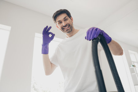 Happy man enjoying weekend cleaning at home with vacuum cleaner and purple gloves in bright interior