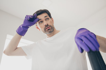 Young man wearing purple gloves taking a break from cleaning in a bright home with thoughtful...