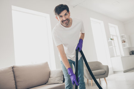 Cheerful man cleaning living room with vacuum in modern home interior on a bright dayの写真素材