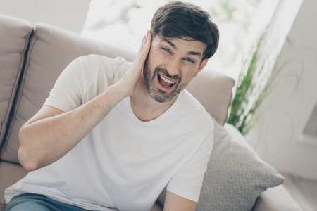 Expressive young man with beard enjoying leisure time on comfortable sofa at home, feel tooth pain in bright daylightの写真素材