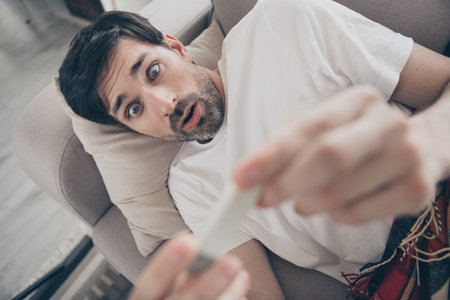 Surprised young man at home discovering unexpected news while relaxing on the sofa during a sunny day indoorsの写真素材
