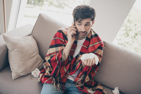 Young man with cold indoors wrapped in plaid blanket talking on phone in cozy home settingの写真素材