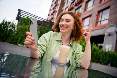Joyful young red-haired woman enjoying coffee while video chatting outdoors on a sunny dayの写真素材