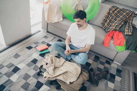 Relaxed young man sitting at home surrounded by clothes and books during a cozy weekend afternoon indoorsの写真素材