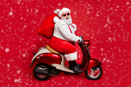 Profile side view portrait of nice bearded cheerful Santa St Nicholas father riding moped hurry up delivering large sack purchase concept isolated on bright vivid shine vibrant red color backgroundの写真素材