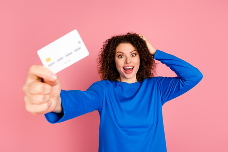 Excited young woman with red hair in blue sweater holding credit card on pink background