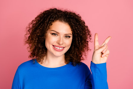 Cheerful young woman making peace sign against a vibrant pink background, wearing a casual blue sweatshirt, showcasing positive vibesの写真素材