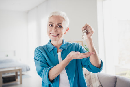 Photo of nice aged lady hold show keys wear blue shirt bright interior apartment indoorsの写真素材