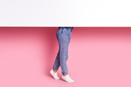 Cropped photo of anonymous woman in denim jeans and white stylish sneakers hiding behind paper wall...
