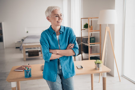 Photo of attractive senior woman crossed hands look window dressed blue outfit cozy day light home interior living roomの写真素材