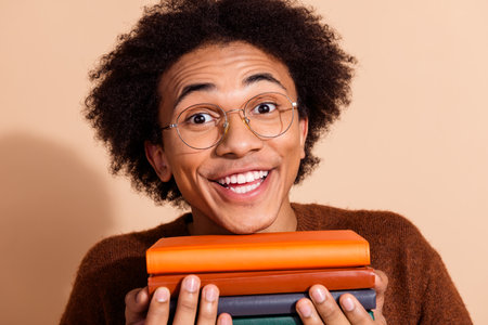 Joyful young student with glasses holding colorful books and beaming smile against beige background.の写真素材