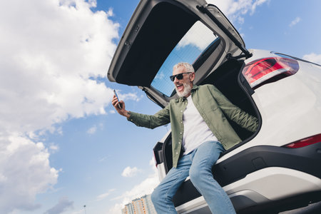 Photo of cheerful positive guy dressed khaki shirt sitting vehicle trunk taking selfie modern device...