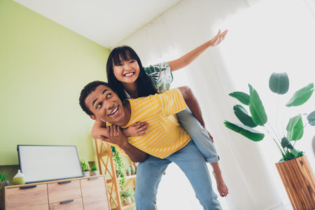 Photo of funny nice multinational couple wear t-shirts have fun time together hold hands piggyback indoors flatの写真素材