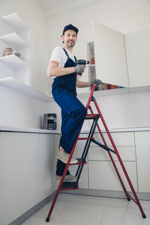 Smiling handyman on a ladder fixing kitchen cabinets during a home renovation projectの写真素材