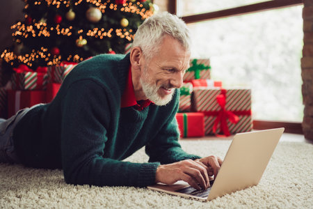 Portrait of nice aged man lying carpet floor use laptop wear sweater new year xmas time flat indoorsの写真素材